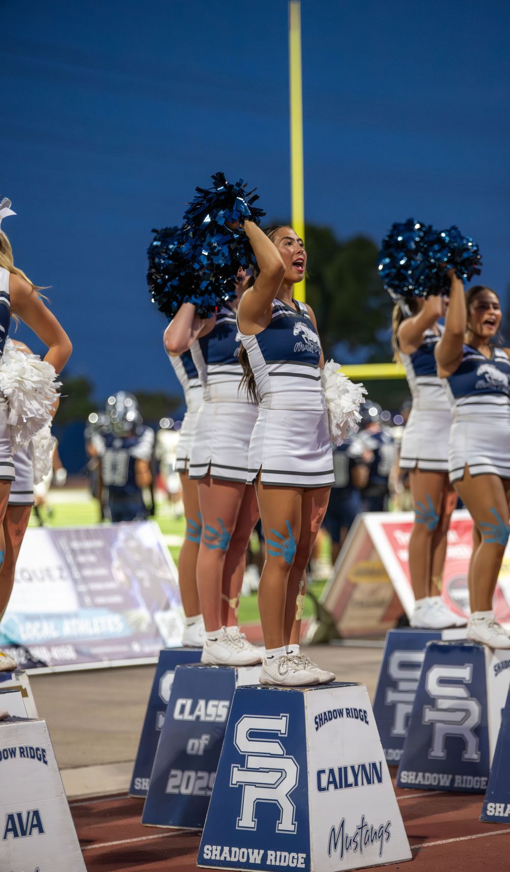 Shadow Ridge Mustangs cheerleaders perform with blue and white pom-poms on the sidelines during a night football game.