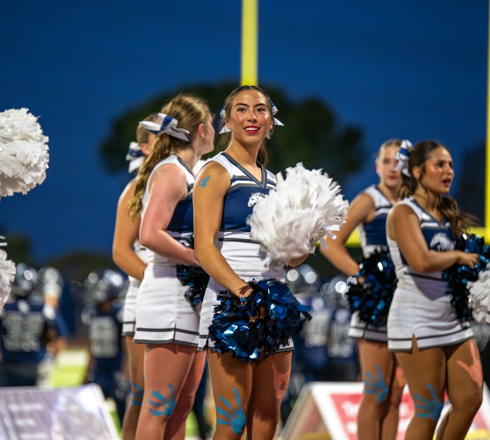 Cheerleaders in blue and white uniforms with pom-poms perform at an outdoor football game.