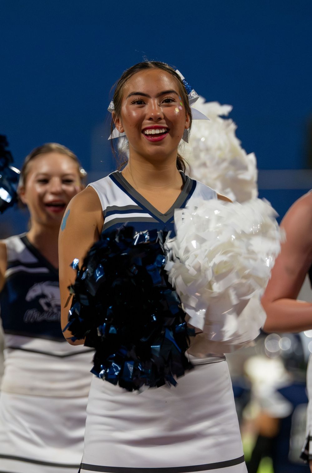 Smiling cheerleader holding black and white pom-poms in a blue and white uniform at a night football game.