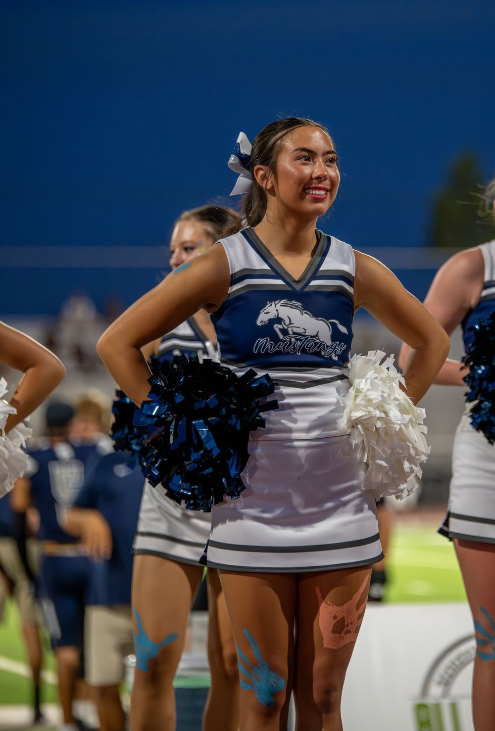Smiling cheerleader in blue and white uniform holds pom-poms at a night football game.