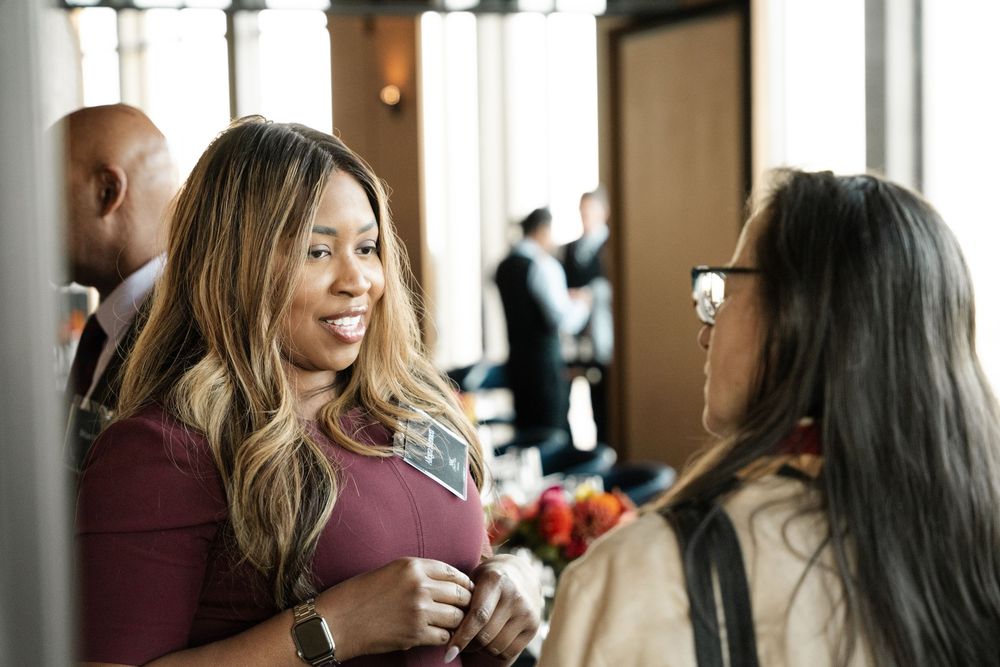 Two professional women networking at a business event, one holding a name badge while engaging in conversation.