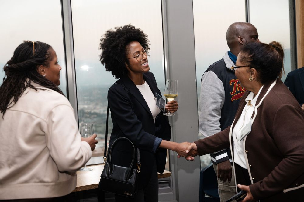 Professionals networking at a high-rise event, shaking hands and socializing near large windows with city views.