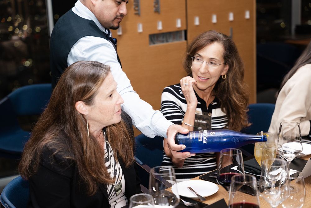 A waiter pours blue bottled water for two women dining at a restaurant table with wine glasses.