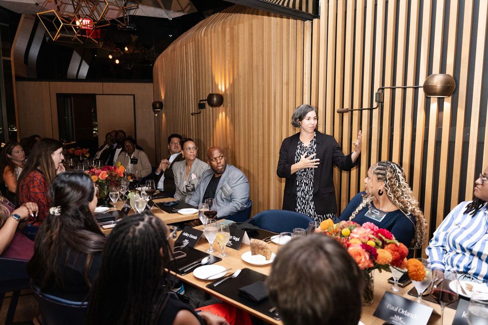 A woman speaks to a diverse group of guests seated at a dinner event in a modern restaurant with warm lighting.