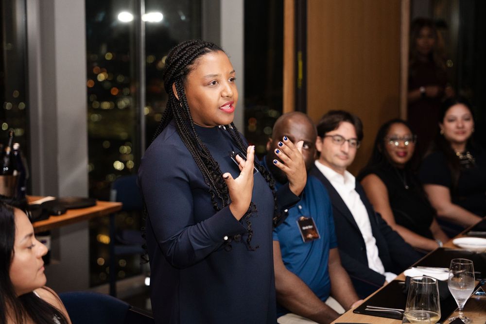 A woman in a navy dress speaks at a dinner event, with attentive guests seated at a restaurant table behind her.