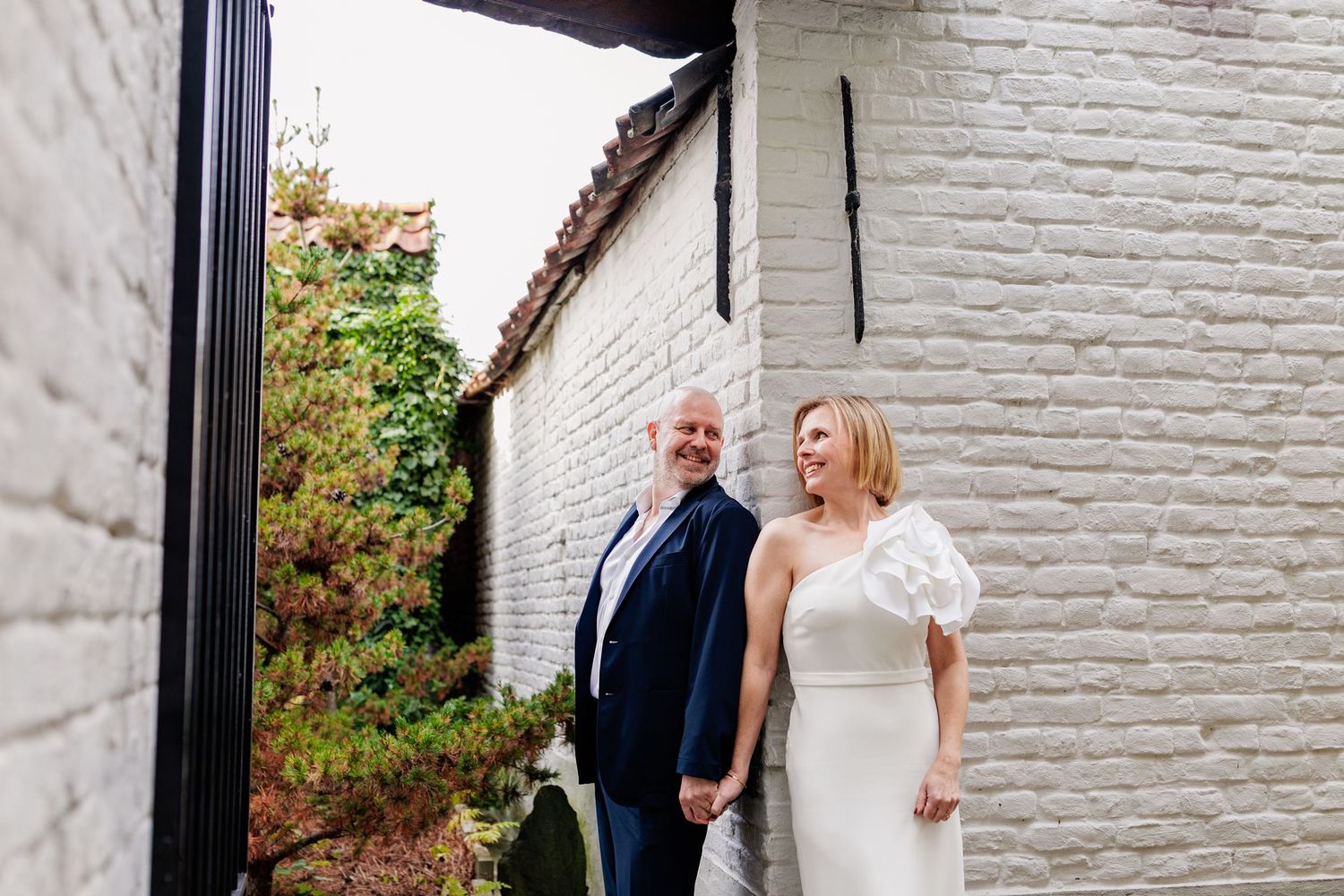 Bride in white gown and groom in navy suit hold hands smiling outdoors beside a white brick wall.