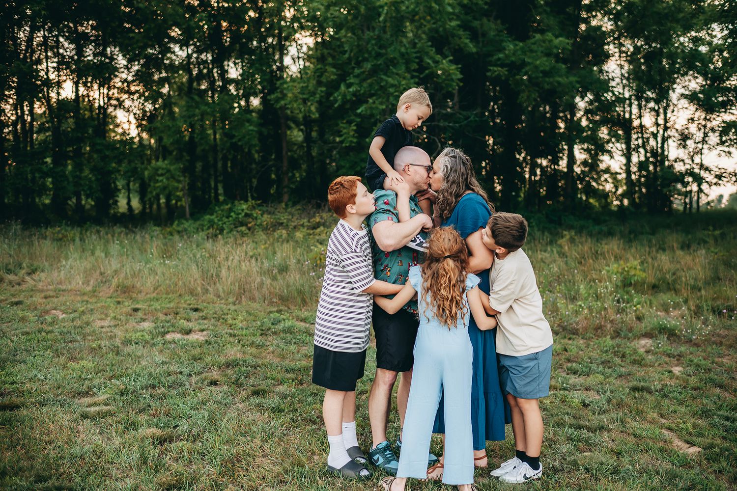 Family group hug in a green meadow at sunset, parents and children embracing outdoors.