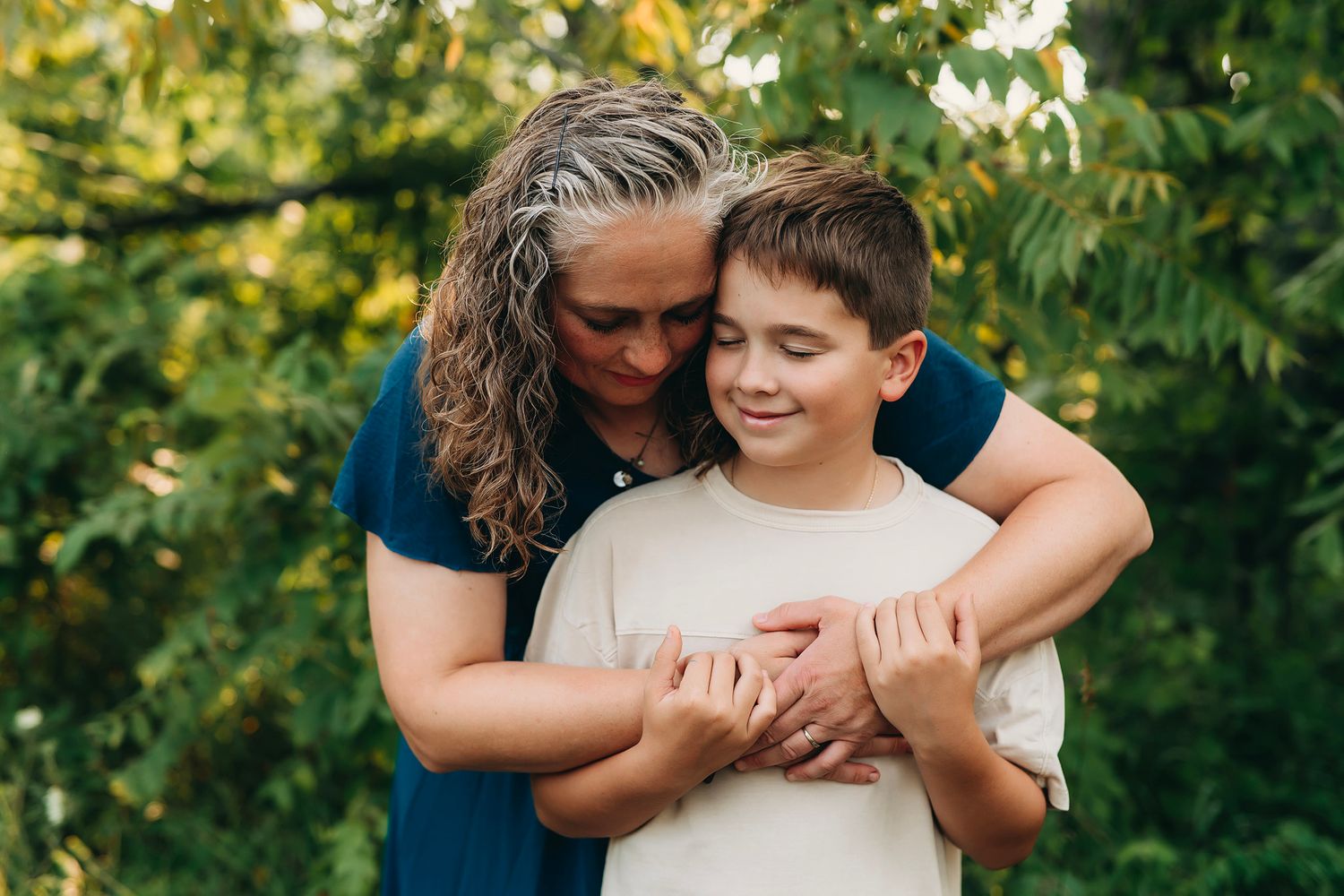 A mother with curly hair tenderly embraces her smiling son outdoors with lush green trees in the background.