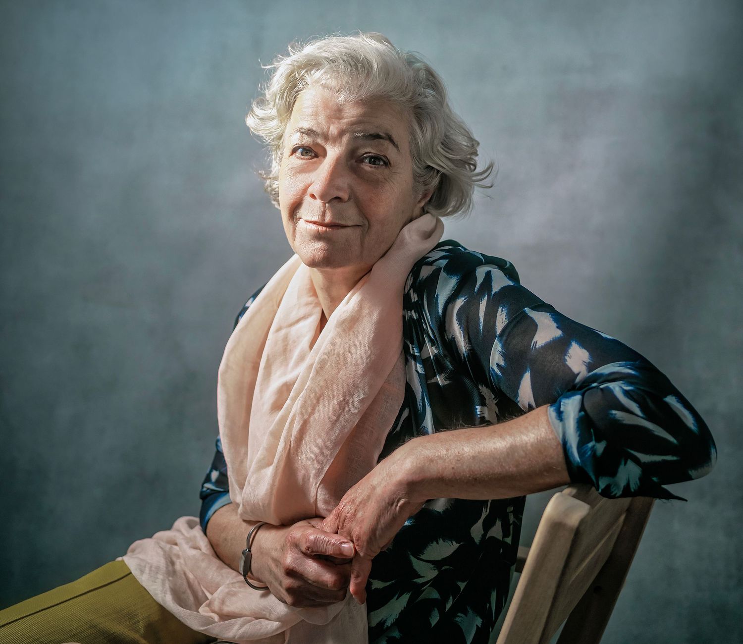 Elderly woman with white hair sits gracefully on a chair, wearing a floral blouse and pink scarf, posing for a portrait.