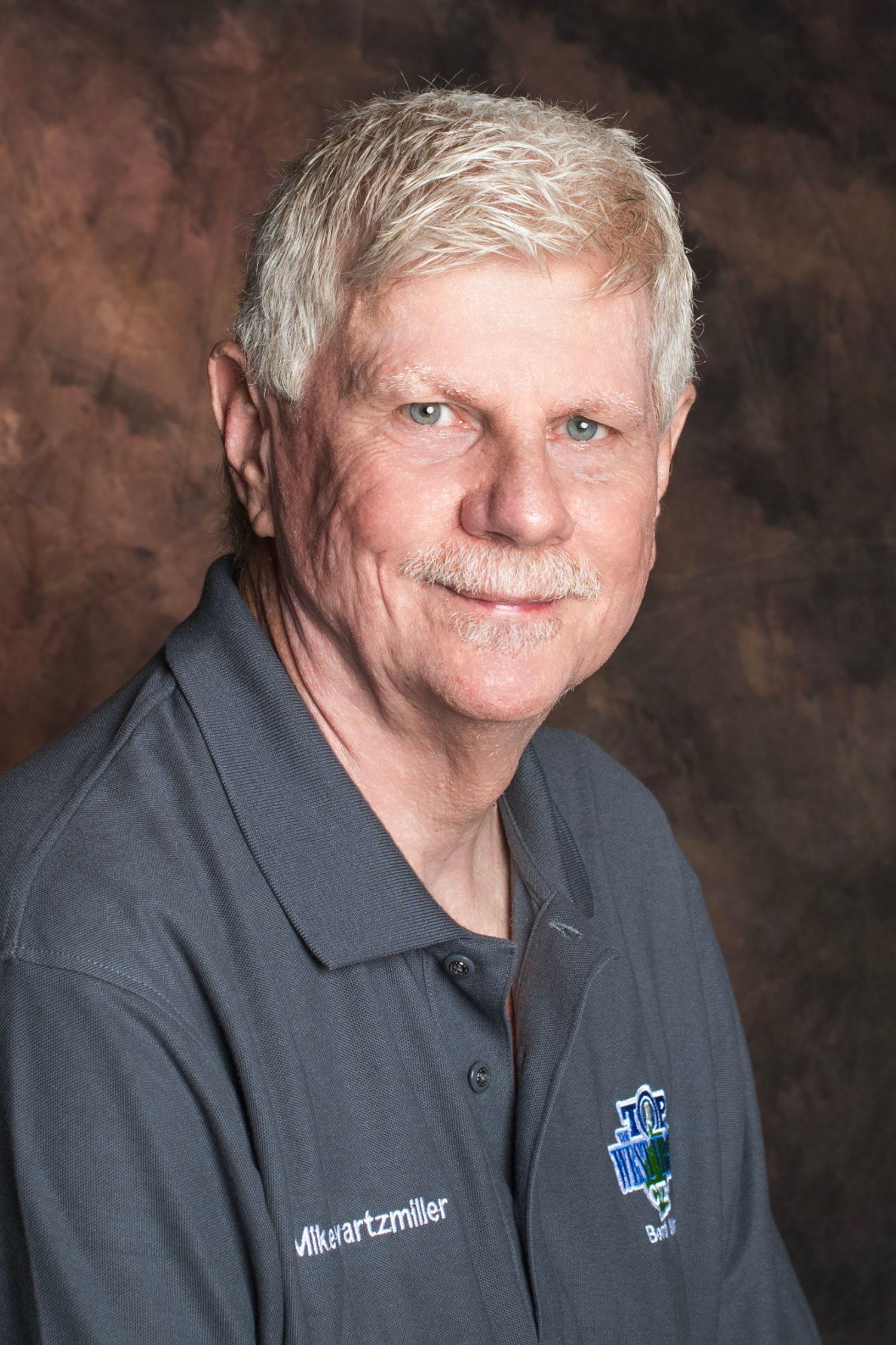 Older man with gray hair and mustache wearing a dark gray Schwartzmiller polo shirt, professional headshot photo.