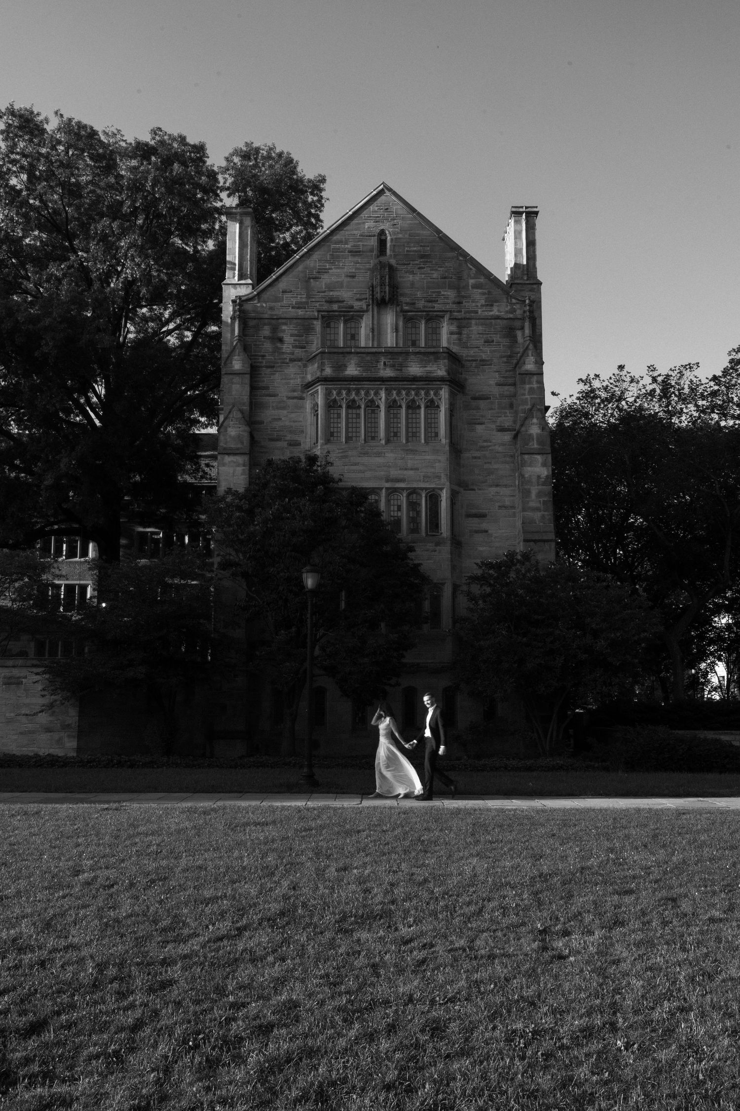 Black and white photo of a couple in wedding attire walking on a path before a grand Gothic stone building at dusk.