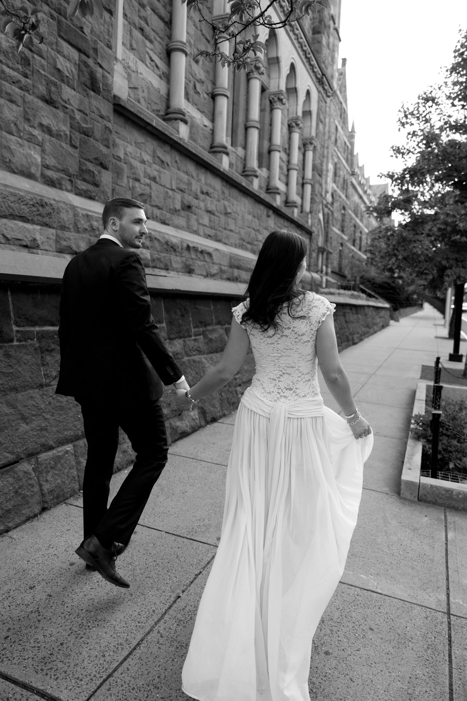 Black and white photo of a bride and groom walking hand in hand along a sidewalk beside a stone building.