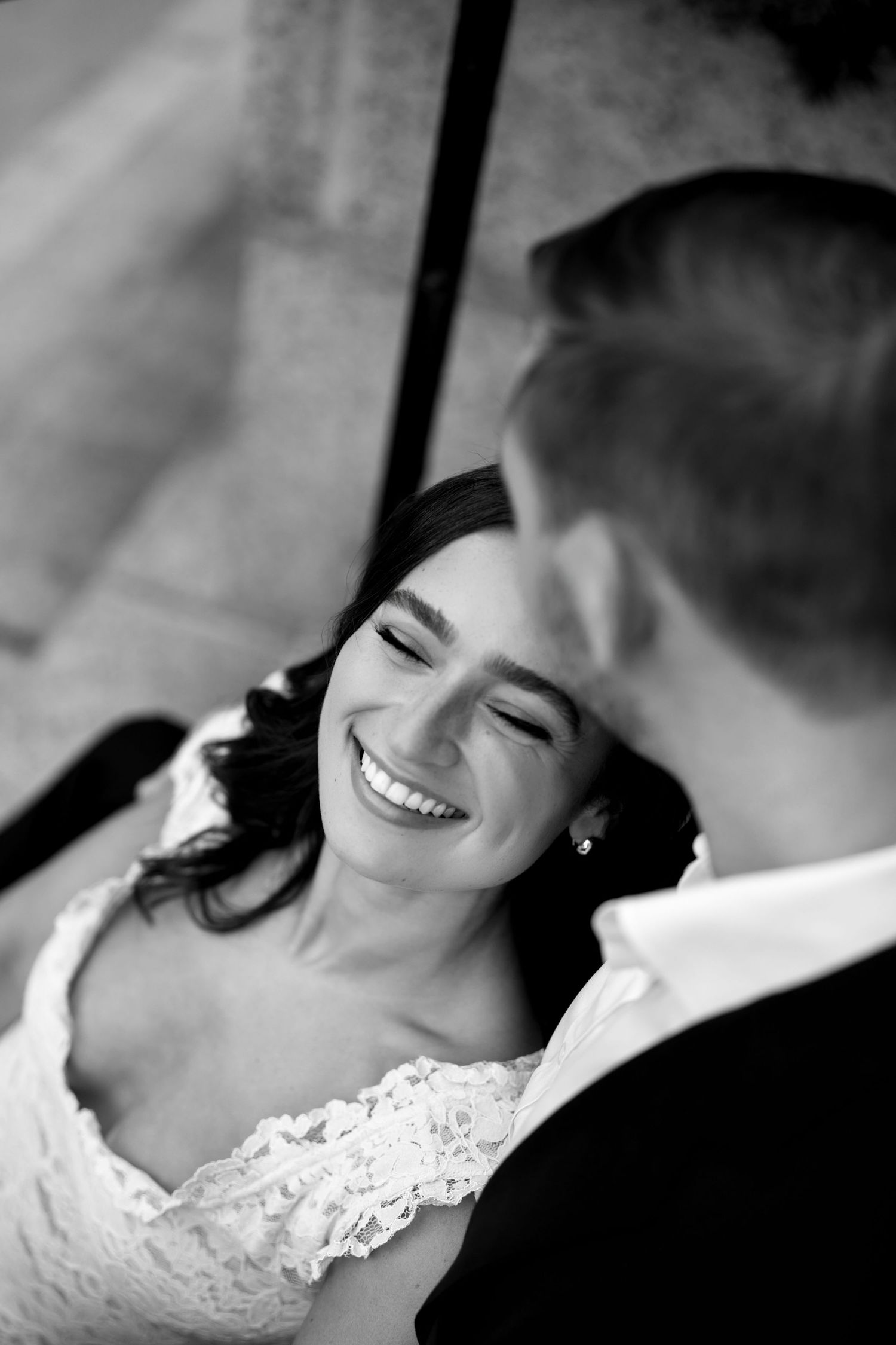 Black and white photo of a joyful bride in lace dress laughing with groom at wedding ceremony.