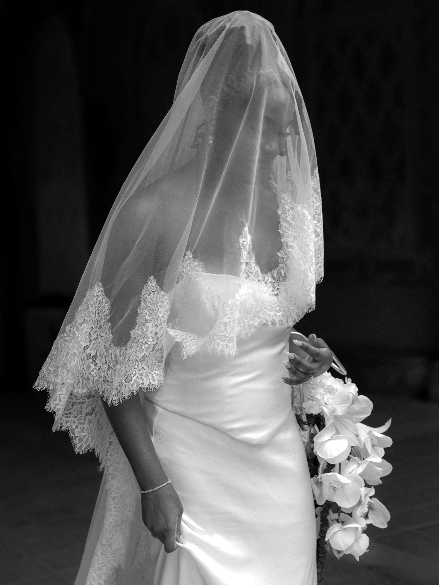 Black and white photo of a bride in a white gown and lace veil holding a bouquet of white flowers.