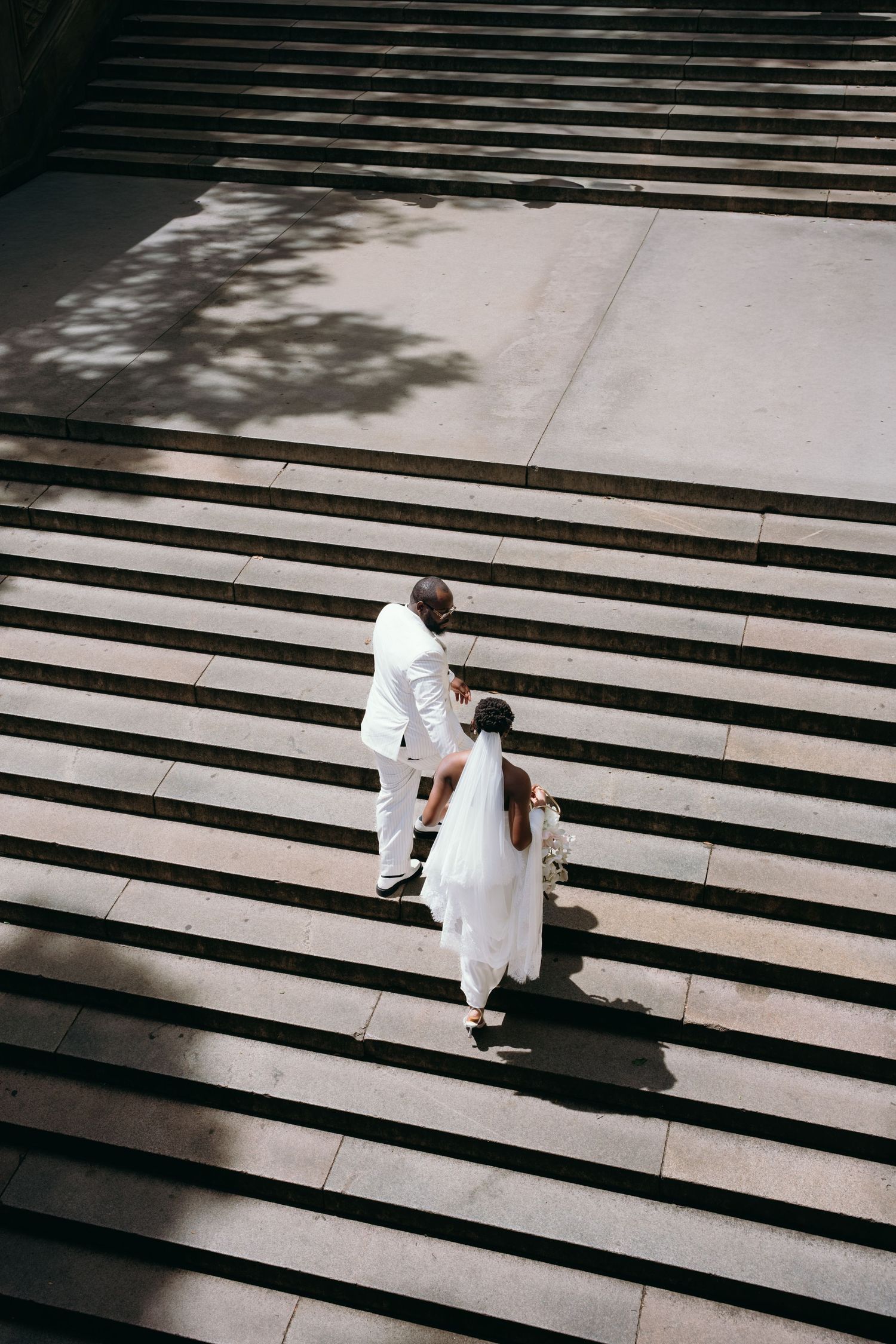 Aerial view of bride and groom in white attire walking up wide stone steps, casting shadows in sunlight.