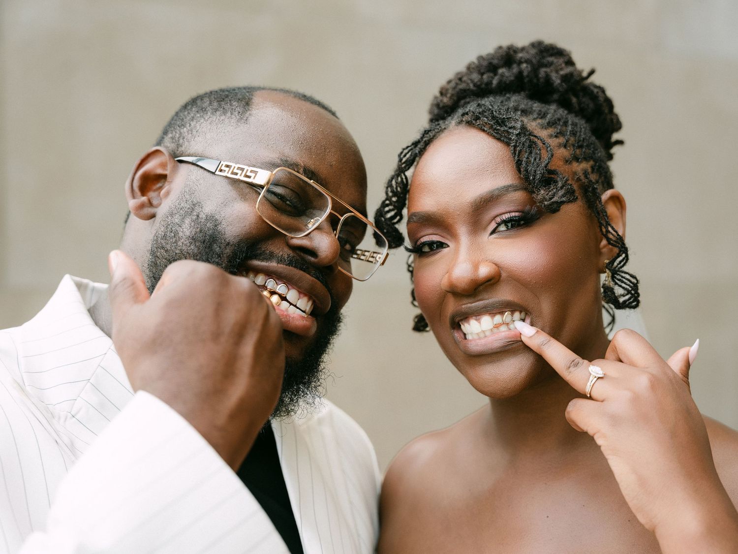 Smiling Black couple in wedding attire, groom in white suit with glasses, bride in white dress showing rings.