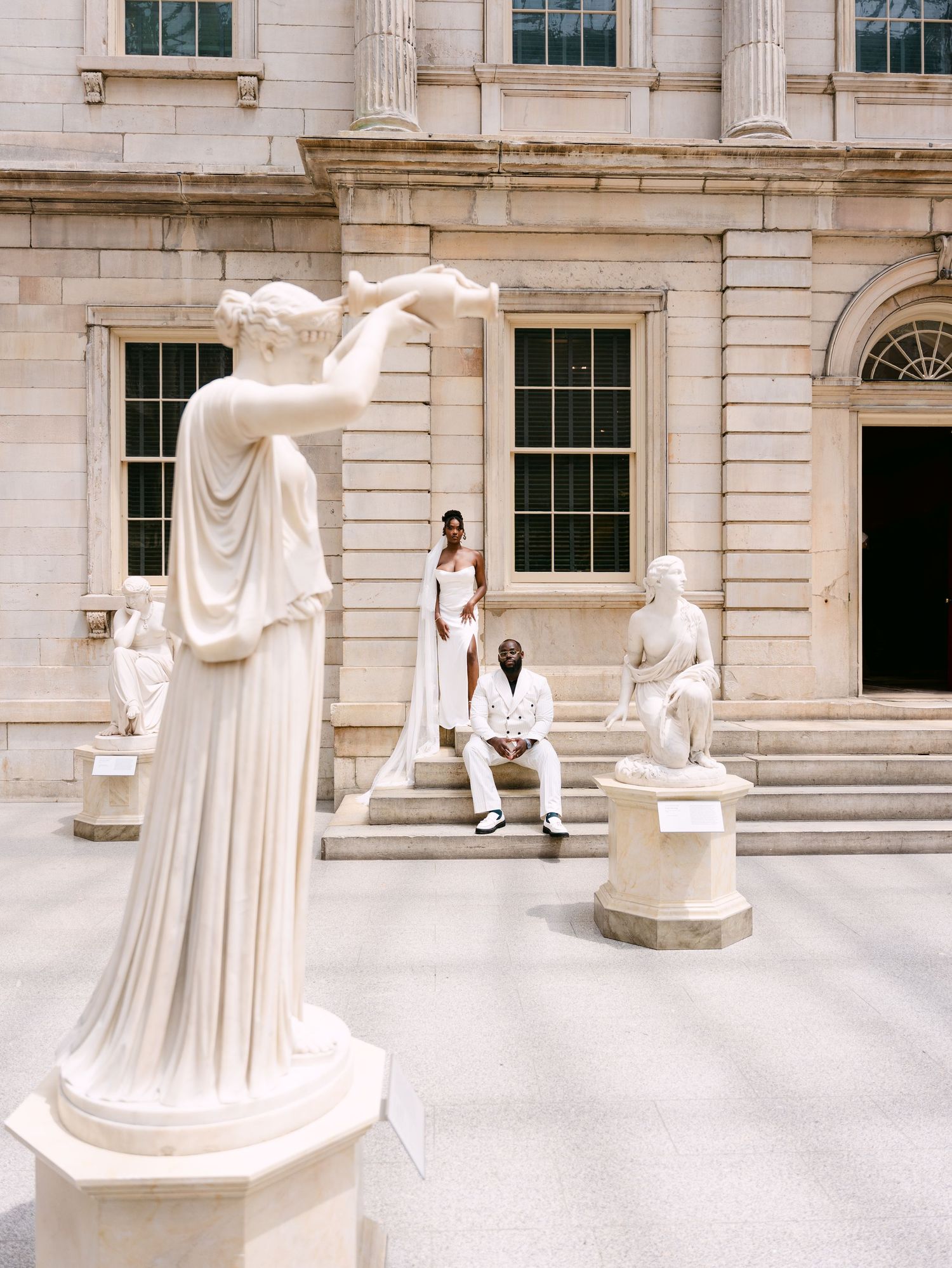 Couple in white attire posing among classical marble statues in a neoclassical building courtyard.