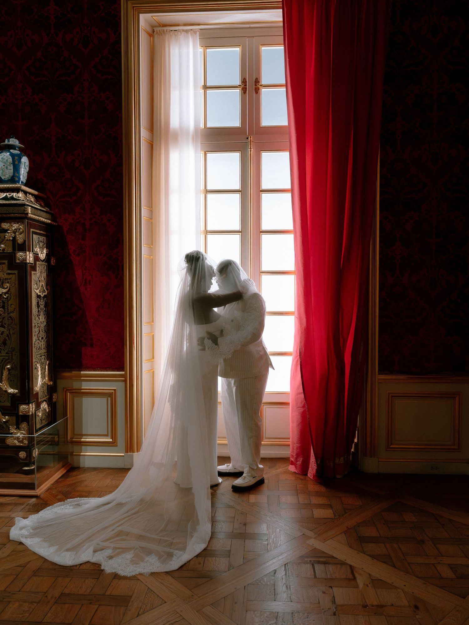 Bride and groom share an intimate moment by a grand window with red curtains in an elegant French chateau.