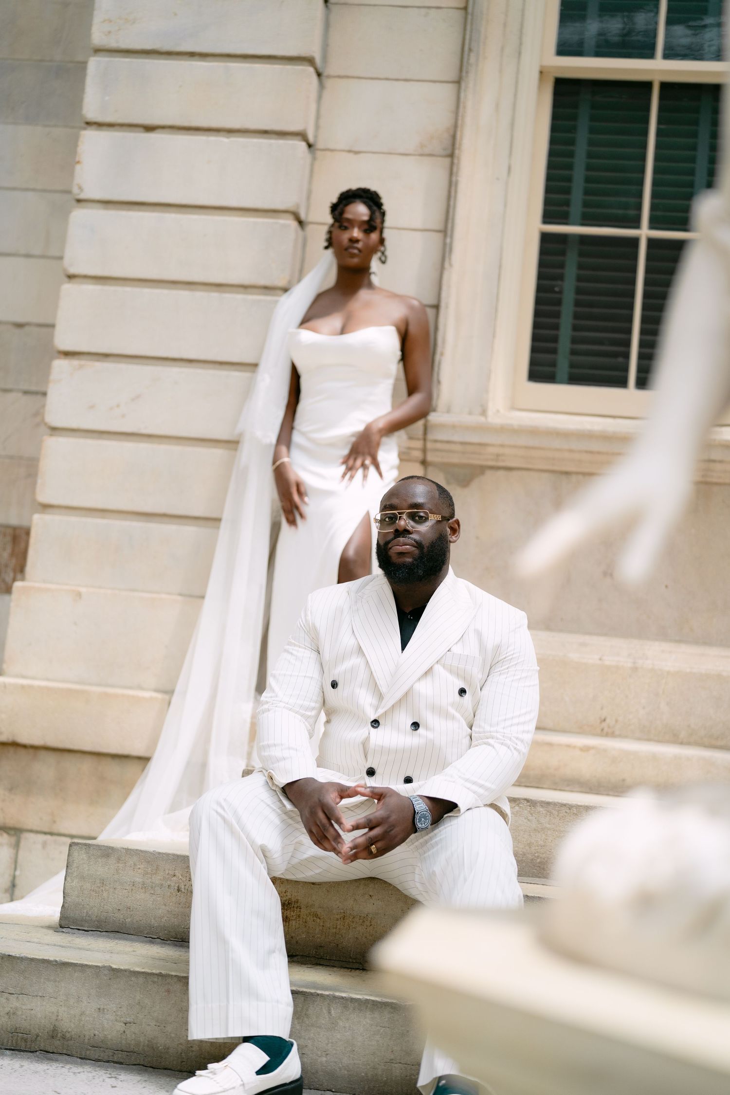A bride in a white strapless gown with cape and groom in white pinstripe suit pose on stone steps.