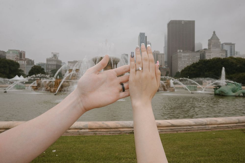 Two hands with rings raised near Buckingham Fountain in Chicago on a cloudy day.
