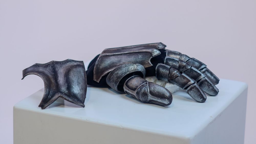 Medieval knight gauntlet sculpture in dark metallic finish displayed on a white pedestal against gray background.