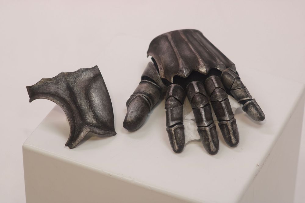 Medieval knight gauntlet armor glove displayed on white pedestal, dark metal articulated finger plates museum exhibit.