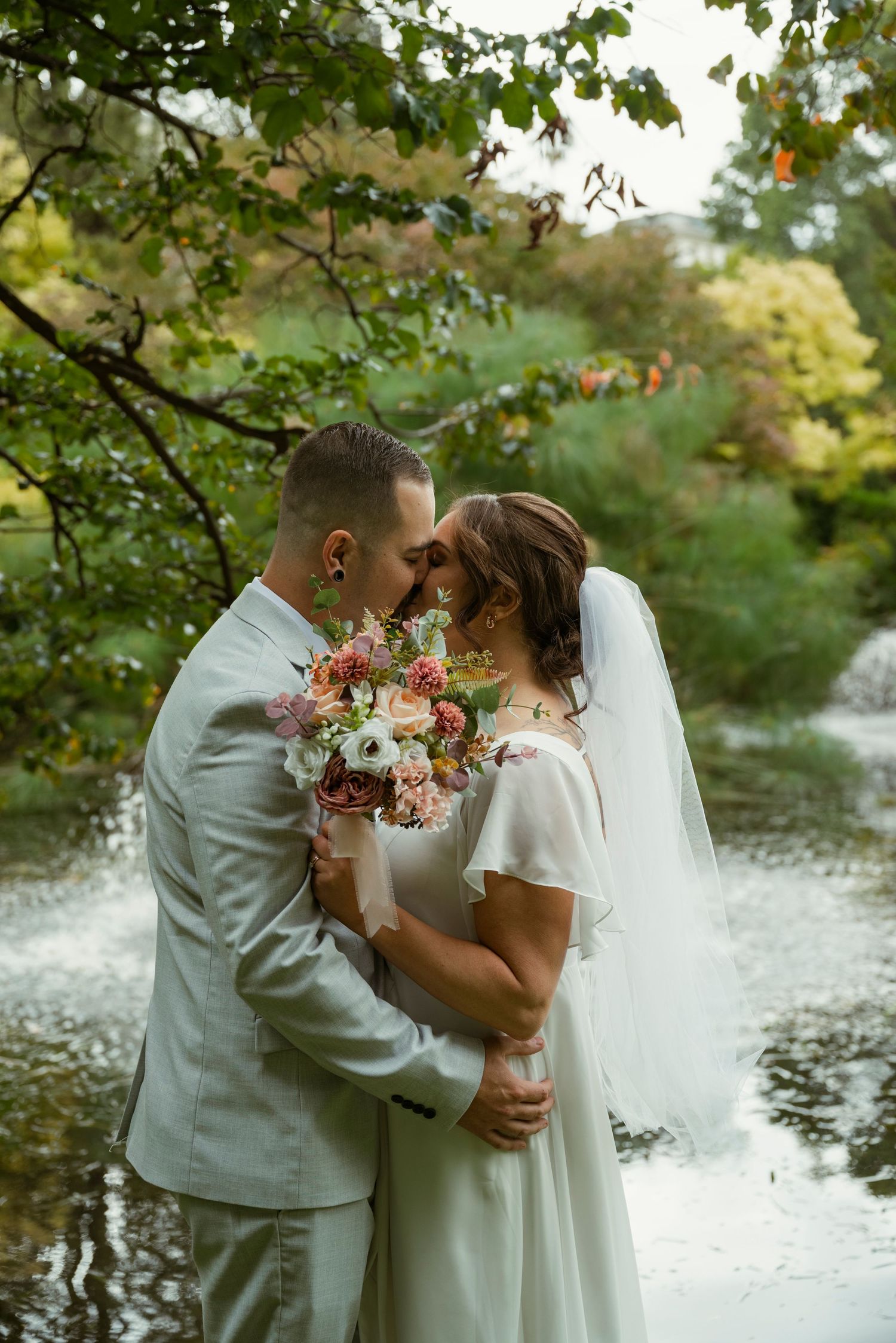 Bride and groom share a romantic moment, touching foreheads while holding a floral bouquet near a serene garden pond.