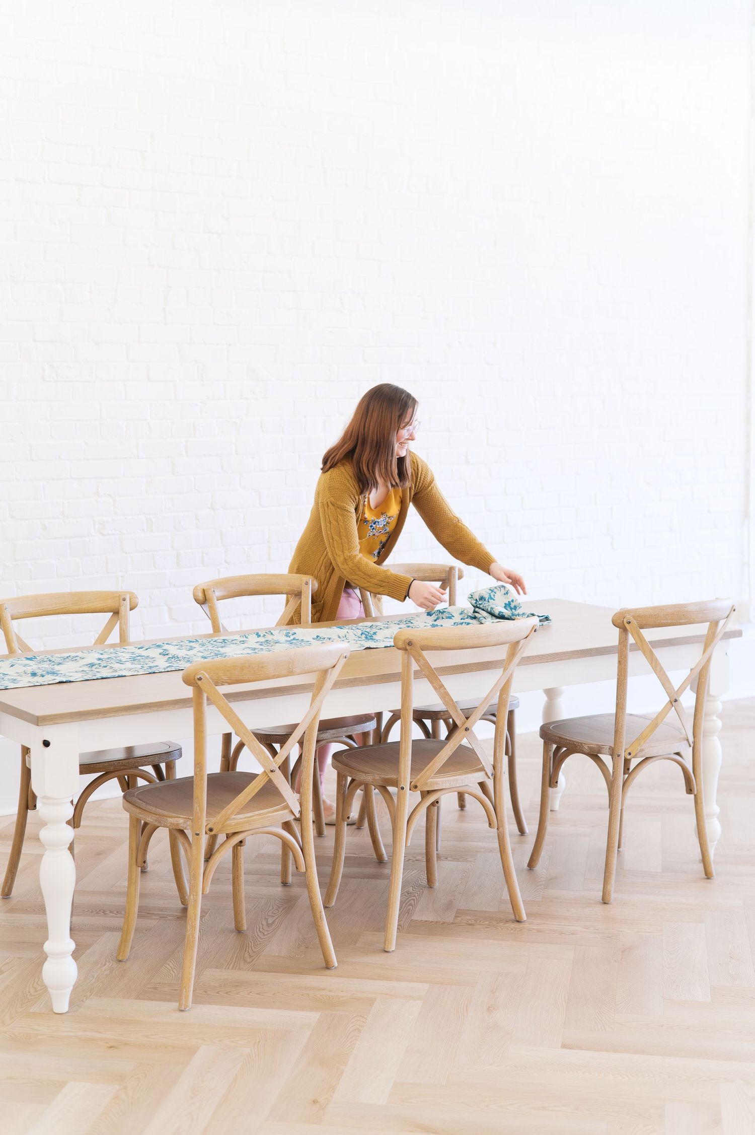 Woman in yellow cardigan arranging a floral table runner on a white farmhouse dining table with natural wood cross-back chairs.