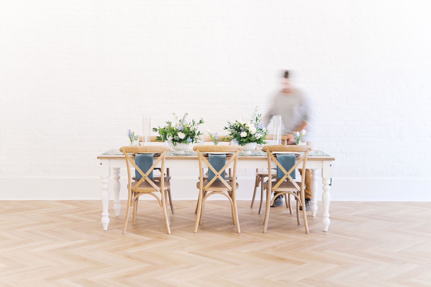 Elegant farmhouse dining table with cross-back chairs, greenery centerpieces, and a blurred figure on herringbone wood floors.