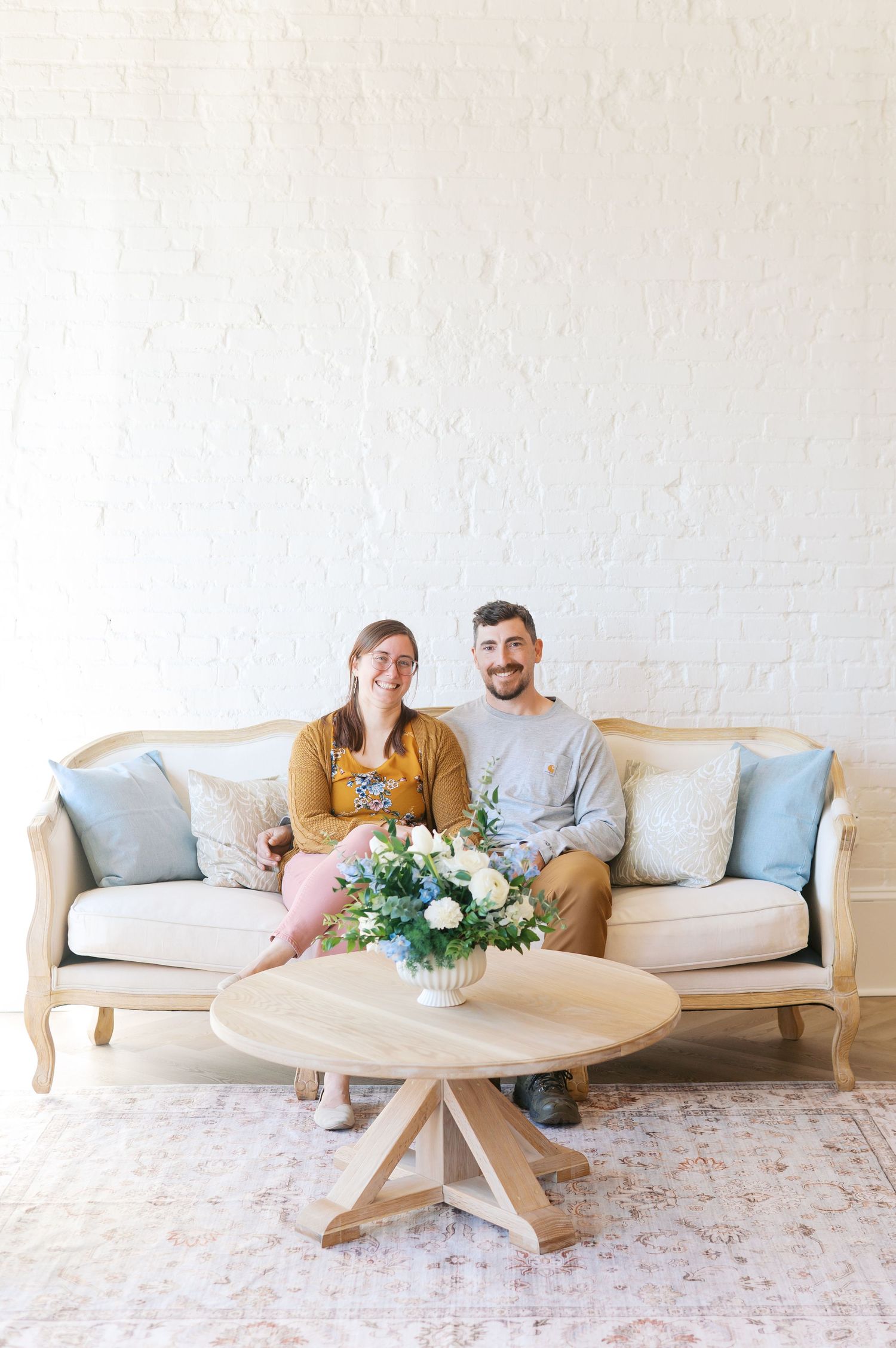Couple sitting together on a vintage beige sofa with blue pillows, smiling, with a floral centerpiece on a round wood table.