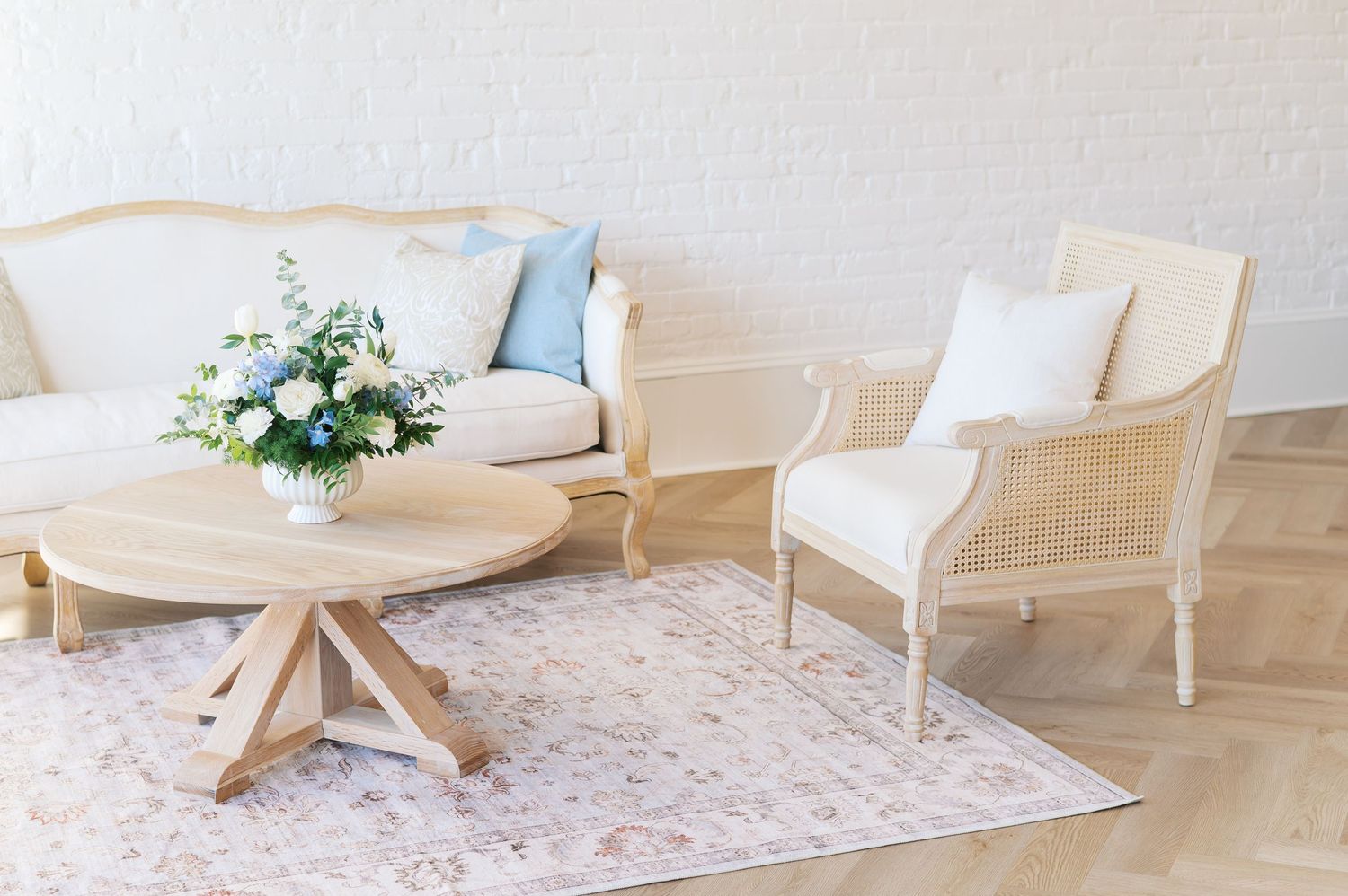 Elegant French-style living room with cream sofa, armchair, round wood coffee table, floral arrangement, and soft pastel rug.