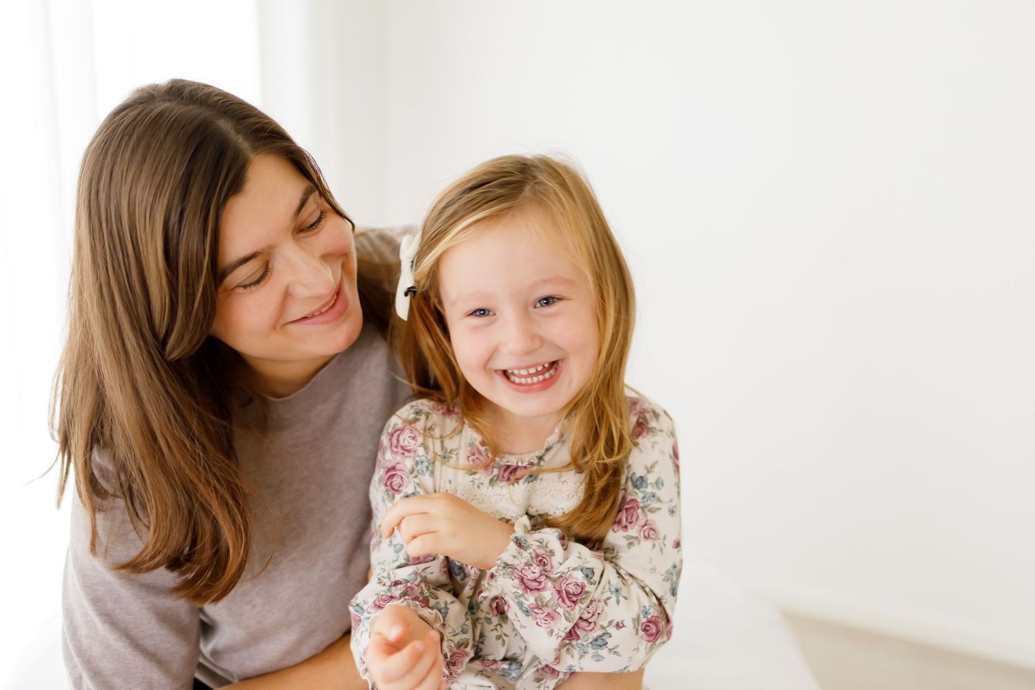 Smiling mother hugging happy young daughter in floral dress against bright white background.