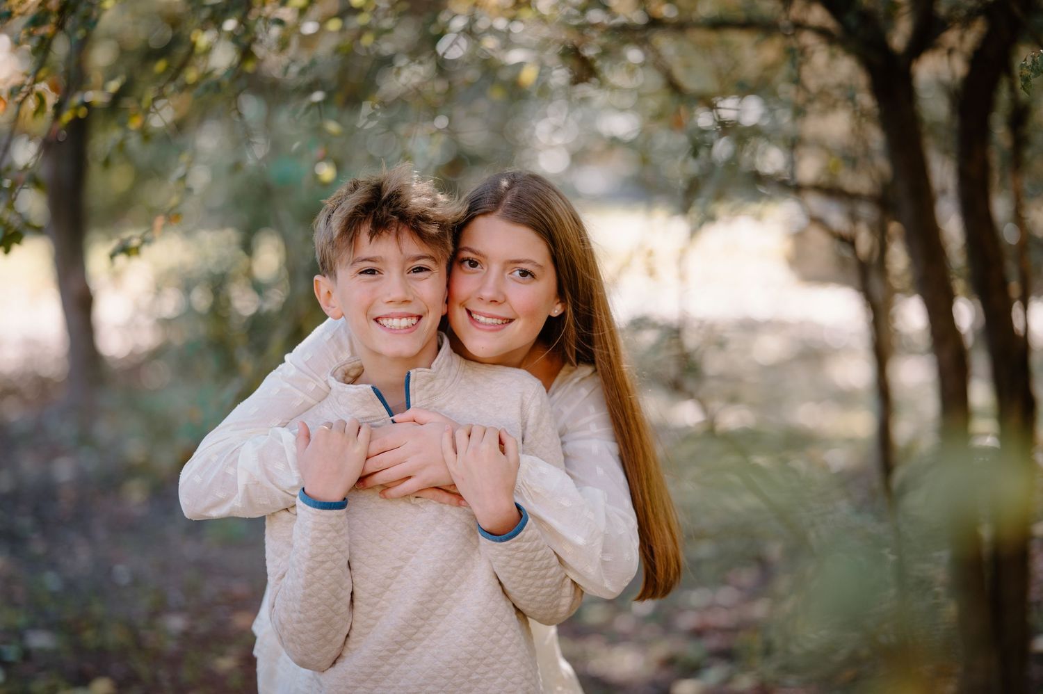 Two smiling children hugging outdoors in a wooded park setting, wearing matching cream sweaters.