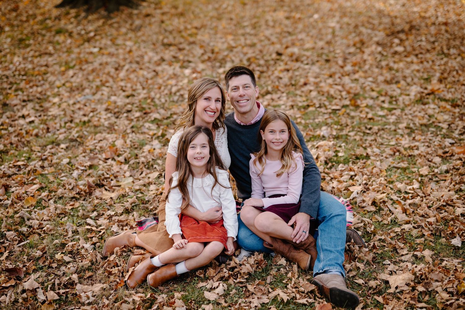 Family of four smiling together on autumn leaves in a park, parents with two young daughters in fall outdoor portrait.