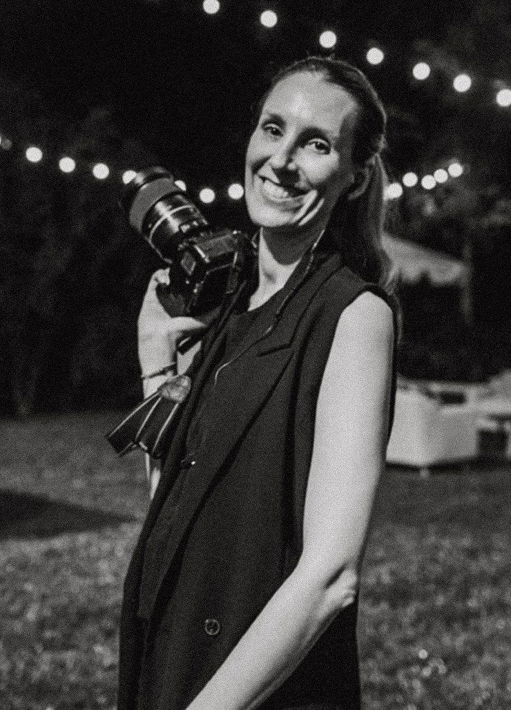 Smiling female photographer holding a camera outdoors at night with string lights in background.