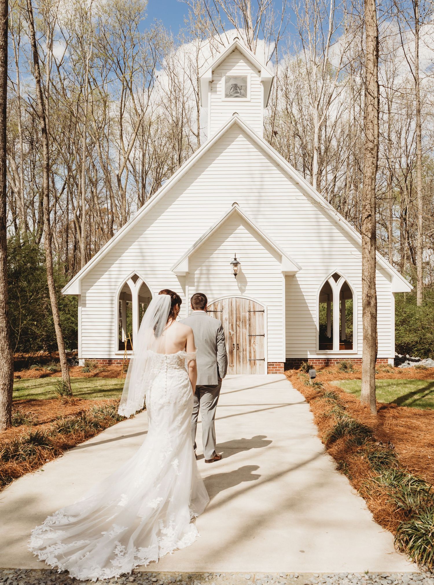 Bride and groom walking toward a charming white chapel surrounded by trees on their wedding day.