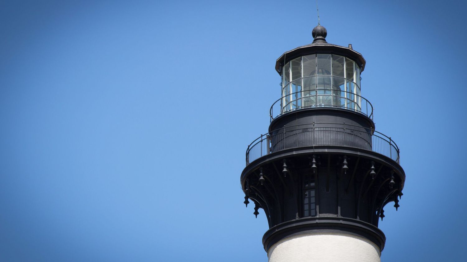 Black and white lighthouse top with glass lantern room against a clear blue sky.