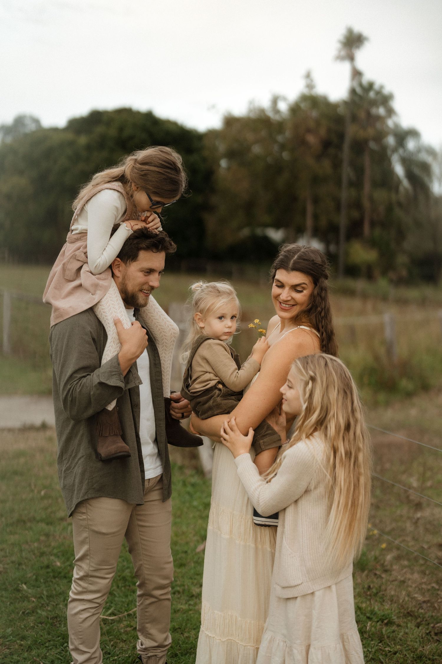 A happy family of five poses outdoors on green grass, with two adults holding three young children near trees.