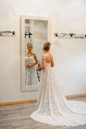 Bride in lace wedding gown with train views her reflection in a full-length mirror inside a bridal boutique.