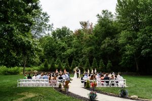 Outdoor wedding ceremony with guests seated on white chairs along a garden path surrounded by lush green trees.