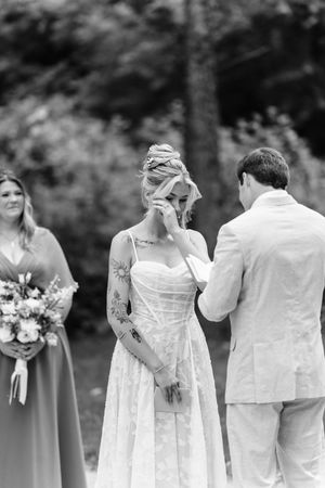 Black and white photo of emotional bride wiping tears during outdoor wedding ceremony with bridesmaid holding bouquet nearby.