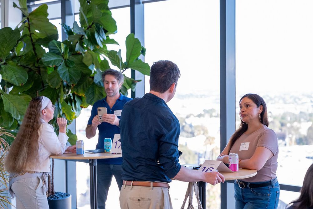 Group of professionals networking at a standing table near large windows with city views.