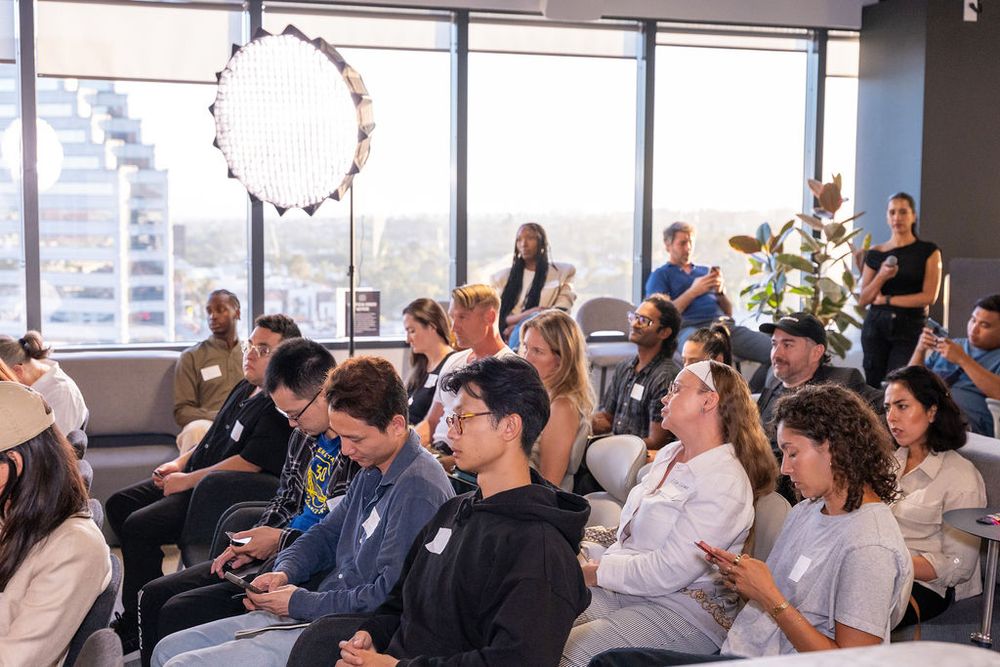 A diverse audience attends an indoor corporate event in a modern office with large windows and bright lighting.