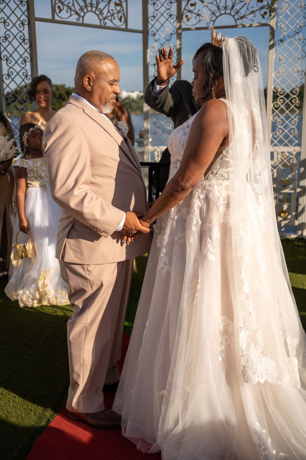 A bride and groom hold hands at an outdoor wedding ceremony, with guests and an officiant visible in the background.
