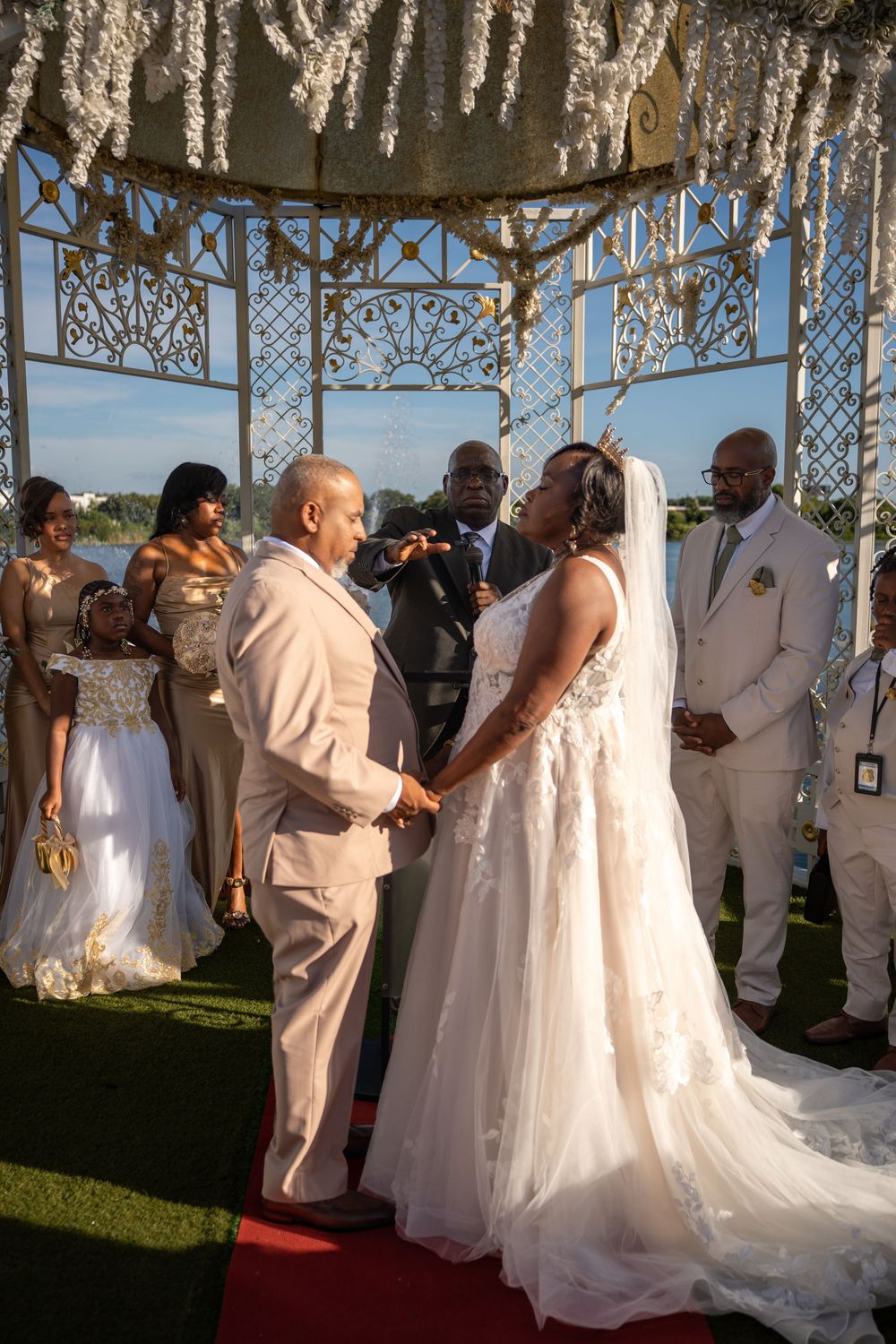 Bride and groom exchange vows under ornate white gazebo during outdoor wedding ceremony with bridal party.