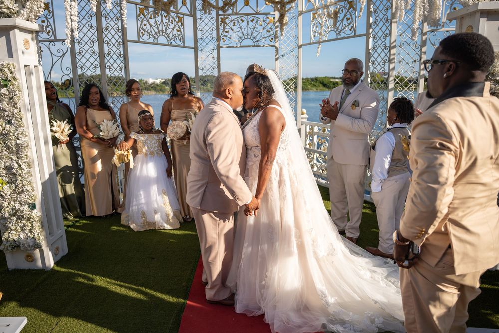 Couple shares first kiss at outdoor waterfront wedding ceremony with bridesmaids and groomsmen under elegant white gazebo.