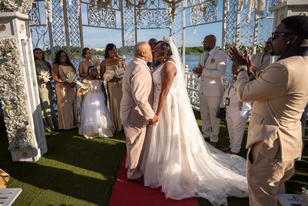 Couple shares a kiss at outdoor wedding ceremony with white gazebo, red carpet, and wedding party in beige attire.