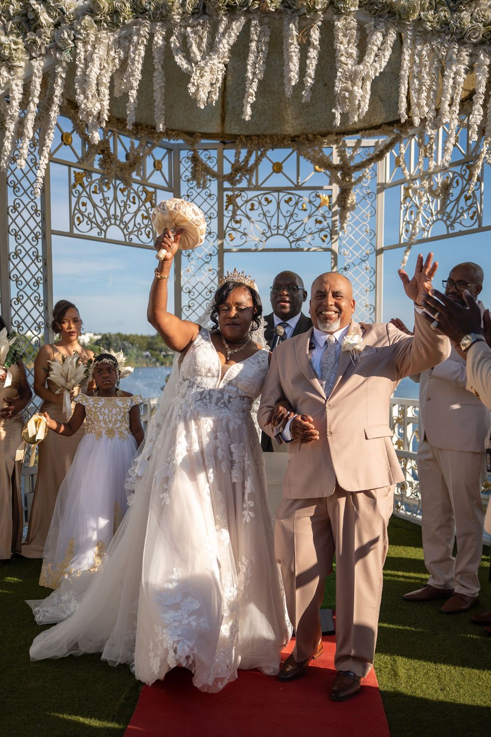 Joyful Black bride in white lace gown and groom in tan suit celebrate after outdoor wedding ceremony under floral gazebo.