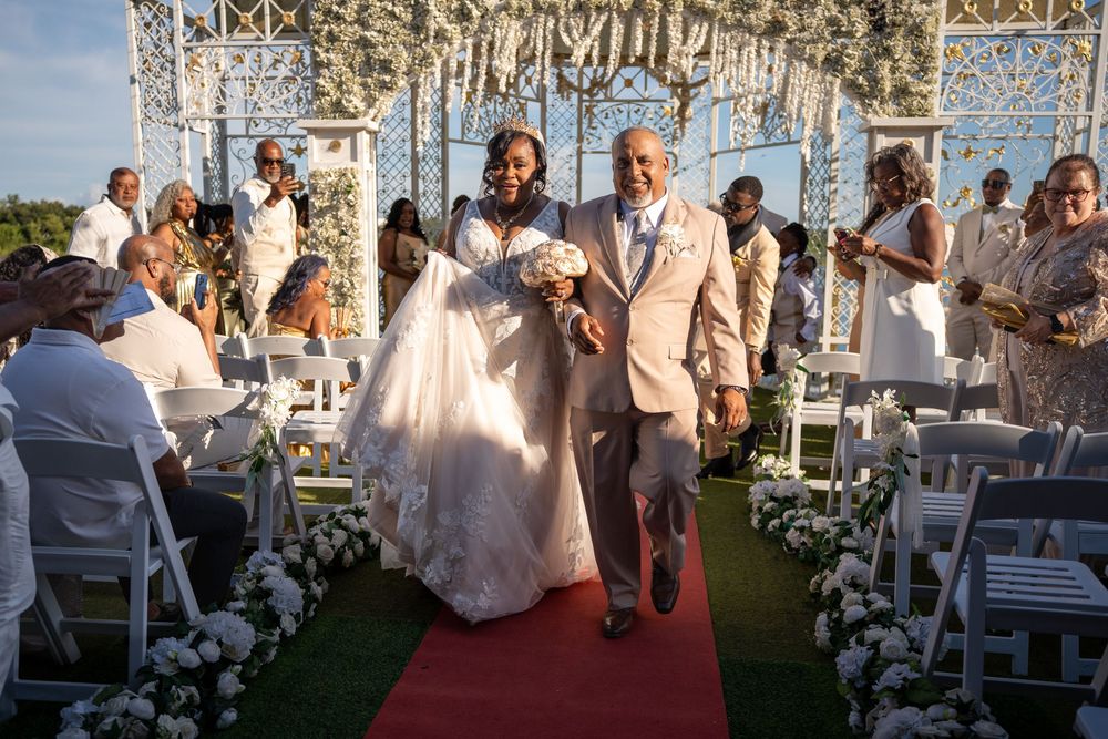Bride and groom walk down red carpet aisle at outdoor wedding ceremony with white floral arch and guests.