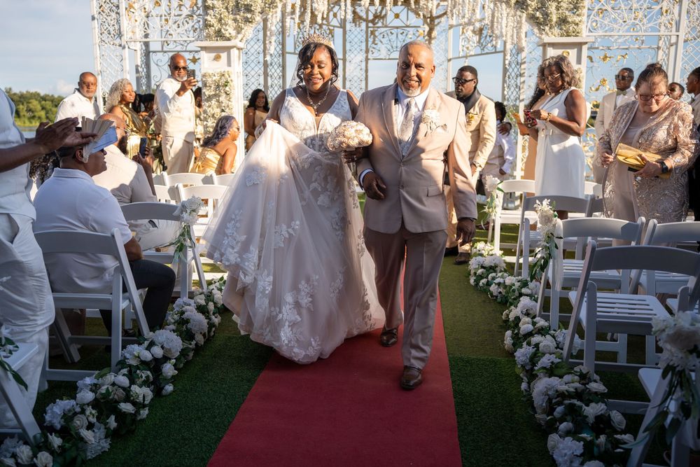 Bride and groom walk down red carpet aisle at outdoor wedding ceremony with white floral decorations.